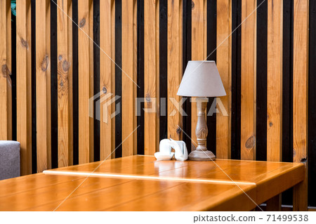 A retro style wooden table lamp stands on an table with napkin by the decorative wall of brown planks and black stripes in the terrace cafe, nobody. A retro style wooden table lamp stands on an table with napkin by the decorative wall of brown planks and black stripes in the terrace cafe, nobody. 71499538