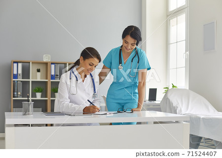 Young smiling women doctor and nurse looking at table at written notes together Young smiling women doctor and nurse looking at table at written notes together 71524407