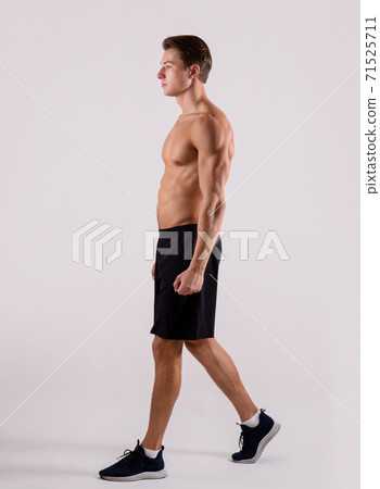 Young sporty guy with sexy bare torso walking on light studio background, side view 71525711