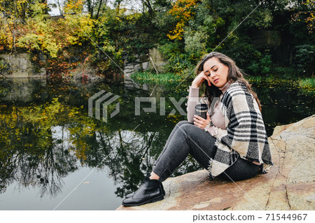 Brunette with long hair sitting next to the lake and drinking hot tea from thermos. Ouutdoor autumn resting. Brunette with long hair sitting next to the lake and drinking hot tea from thermos. Ouutdoor autumn resting. 71544967