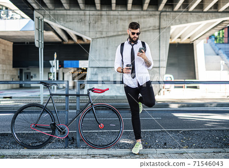 Young man commuter with bicycle standing outdoors in city, using smartphone. 71546043