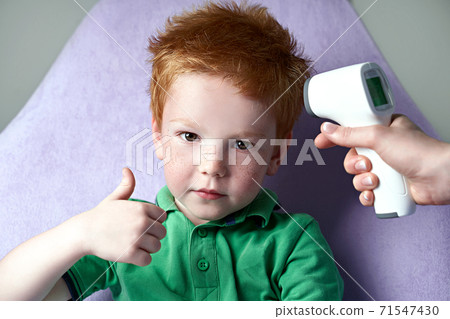 Doctor measures temperature of a cute red haired freckled little boy showing thumb up with a non-contact infrared thermometer. Good health and normal temperature concept. 71547430