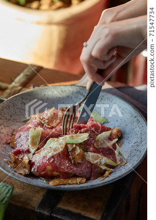 A female hand is preparing a vegetable salad with beef roast beef. A plate of salad with beef roast beef, onions, arugula, pepper and tomatoes on a black table. Top view. 71547734
