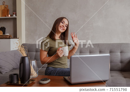 Portrait of pleasant young woman with mug looking at camera, waving hello. Pretty millennial lady communicating with parents via video call 71551798
