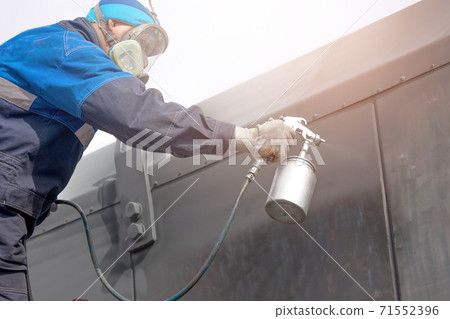 Industrial work. Priming of metal products from the compressor gun. A worker in overalls and a protective mask paints the body of a truck trailer or a metal car. 71552396