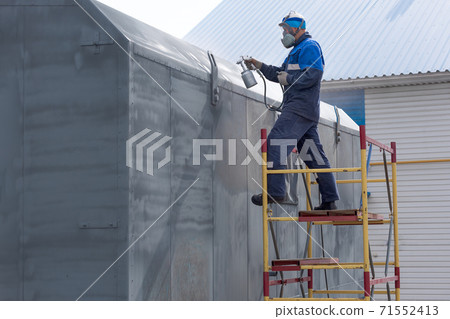 Industrial work. Priming of metal products from the compressor gun. A worker in overalls and a respirator paints the body of a truck trailer or a metal car. Industrial work. Priming of metal products from the compressor gun. A worker in overalls and a respirator paints the body of a truck trailer or a metal car. 71552413