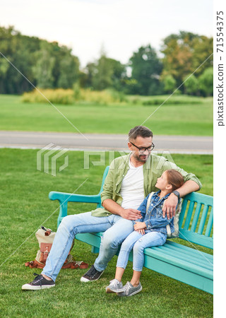 Full lentgh shot of happy young father and his cute little daughter talking while sitting on the bench, spending time together in the green park Full lentgh shot of happy young father and his cute little daughter talking while sitting on the bench, spending time together in the green park 71554375
