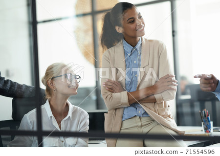 Two smiling diverse female coworkers looking aside while having a meeting with colleagues in the modern office 71554596