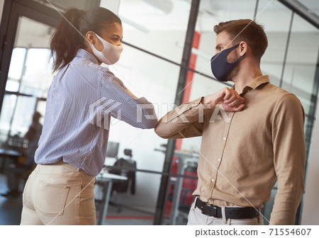 Two young diverse business colleagues wearing face protective masks bumping elbows, greeting each other while working during covid 19 quarantine 71554607