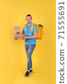 Young smiling guy delivering food from restaurant in blue uniform with thermo bag on his back holds an order for customer on yellow studio background. Fast food delivery to office or home 71555691