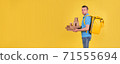 Young smiling guy delivering food from restaurant in blue uniform with thermo bag on his back holds an order for customer on yellow studio background. Fast food delivery to office or home 71555694
