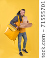 Cute caucasian food delivery courier smiles and looks ahead, holding food order in his hands and yellow refrigerator bag on yellow background hangs on his shoulder. Food delivery to home or office 71555701
