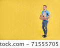 Young smiling guy delivering food from restaurant in blue uniform with thermo bag on his back holds an order for customer on yellow studio background. Fast food delivery to office or home 71555705
