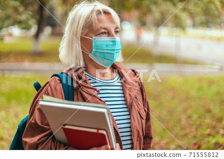 Teacher in casual clothes, glasses and a backpack holding an exercise book, notebook. Blond woman wearing respiratory face mask during coronavirus pandemic 71555812