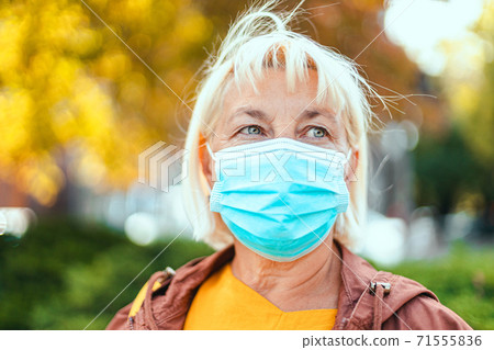 Caucasian adult blond woman 50 years old in bright robe and protective medical respirator mask looking around on the park background 71555836