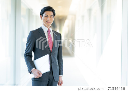Portrait of a young man standing with a computer in the office corridor Portrait of a young man standing with a computer in the office corridor 71556436