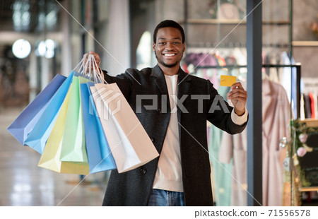 Cheerful millennial african american man posing with credit card and colorful bags with purchases in mall 71556578