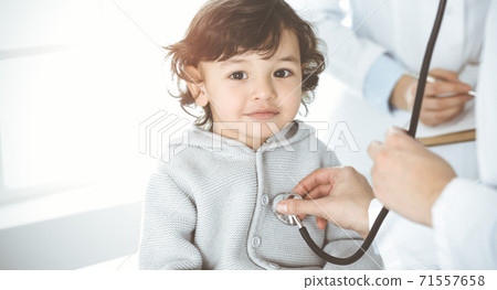 Woman-doctor examining a child patient by stethoscope in sunny clinik. Cute arab toddler at physician appointment Woman-doctor examining a child patient by stethoscope in sunny clinik. Cute arab toddler at physician appointment 71557658
