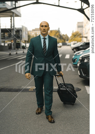 Businessman goes ahead holding suitcase in hand. Handsome Caucasian man walking past hotel parked cars. Business trip concept Businessman goes ahead holding suitcase in hand. Handsome Caucasian man walking past hotel parked cars. Business trip concept 71558096