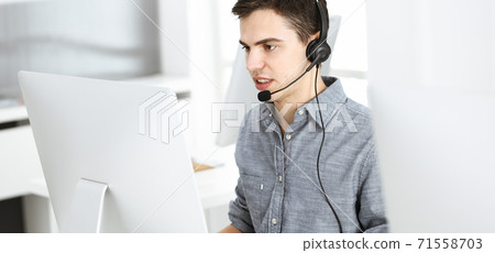 Casual dressed young man using headset and computer while talking with customers online. Call center, business concept Casual dressed young man using headset and computer while talking with customers online. Call center, business concept 71558703