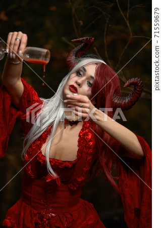 Maleficent Woman in Red Clothing and Horns in dark Forest. Posing with wineglass. Maleficent Woman in Red Clothing and Horns in dark Forest. Posing with wineglass. 71559679