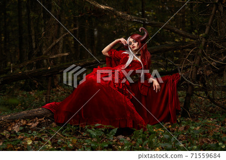 Maleficent Woman in Red Clothing and Horns in dark Forest. Posing in magik forest Maleficent Woman in Red Clothing and Horns in dark Forest. Posing in magik forest 71559684