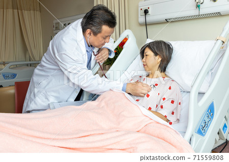 Asian medical doctor examining female patient with stethoscope whom is seated in bed in hospital 71559808