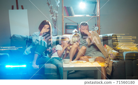 Happy family watching projector, TV, movies with popcorn in the evening at home. Mother, father and kids spending time together. 71560180