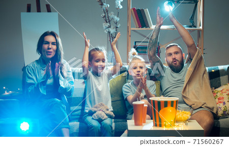 Happy family watching projector, TV, movies with popcorn in the evening at home. Mother, father and kids spending time together. 71560267