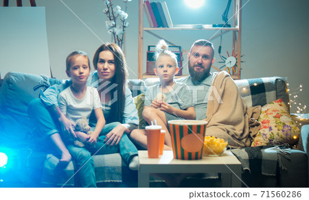 Happy family watching projector, TV, movies with popcorn in the evening at home. Mother, father and kids spending time together. Happy family watching projector, TV, movies with popcorn in the evening at home. Mother, father and kids spending time together. 71560286