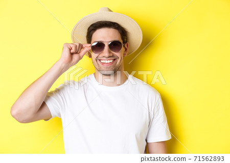 Concept of tourism and vacation. Close-up of handsome man tourist looking happy, wearing sunglasses and summer hat, standing over yellow background Concept of tourism and vacation. Close-up of handsome man tourist looking happy, wearing sunglasses and summer hat, standing over yellow background 71562893
