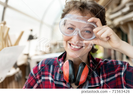 Young beautiful handy professional happy female strong carpenter portrait wearing protective goggles working in carpentry diy workshop against wood. Confident engineer. Women male hobby at workbench 71563426
