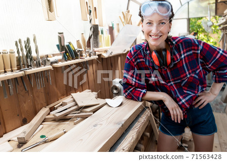 Young beautiful handy professional happy female strong carpenter portrait wearing protective goggles working in carpentry diy workshop against wood. Confident engineer. Women male hobby at workbench 71563428
