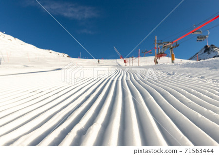 Close-up straight line rows of freshly prepared groomed ski slope piste with bright shining sun and clear blue sky background. Snowcapped mountain downhill landscape at europe winter skiing resort 71563444