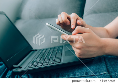 Closeup view of young woman hands working remotely from home, using both laptop and smartphone. Shopping online using mobile app and search engines. Online education and WFH concept. Selective focus Closeup view of young woman hands working remotely from home, using both laptop and smartphone. Shopping online using mobile app and search engines. Online education and WFH concept. Selective focus 71567206