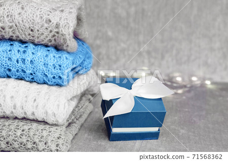 Stylish knitted pastel colored sweaters folded in stack and trendy blue colored present box with white satin bow on velvety fabric background. Winter and spring season knitwear clothing. Color of the Stylish knitted pastel colored sweaters folded in stack and trendy blue colored present box with white satin bow on velvety fabric background. Winter and spring season knitwear clothing. Color of the 71568362