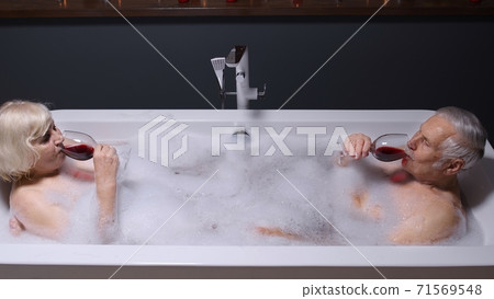 Senior couple man and woman lying in warm bath with bubbles, enjoying relaxation, drinking red wine Senior couple man and woman lying in warm bath with bubbles, enjoying relaxation, drinking red wine 71569548