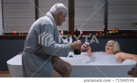 Senior grandmother is taking foamy bath with candles. Grandfather giving glass with champagne 71569559