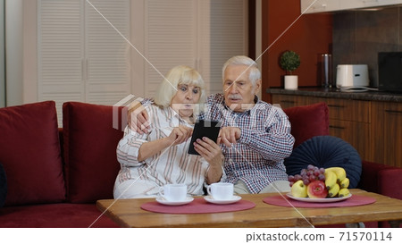 Senior pensioner couple with digital tablet pc computer at home. Resting on sofa in cozy living room Senior pensioner couple with digital tablet pc computer at home. Resting on sofa in cozy living room 71570114