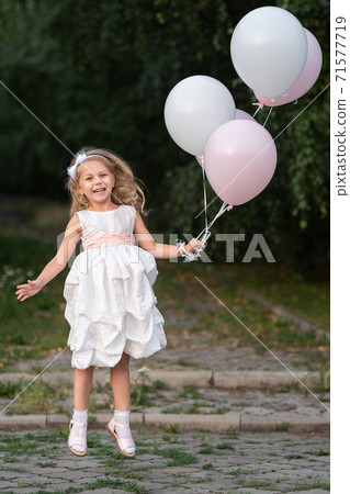 Little girl in white dress with balloons jumping on the street 71577719