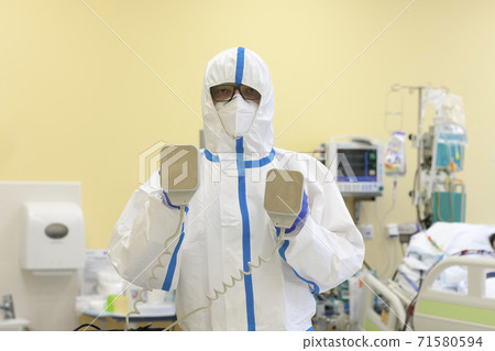 Male nurse with protective coverall clothing in intensive care unit. Nurse is holding defibrillator. The nurse caring for a patient with covid-19. Male nurse with protective coverall clothing in intensive care unit. Nurse is holding defibrillator. The nurse caring for a patient with covid-19. 71580594