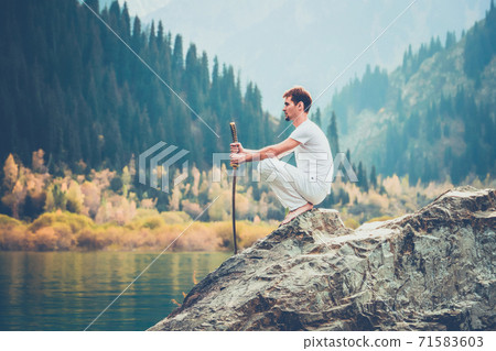A wise man sits on a stone and holds a sword in his hands. Mountain Lake background. 71583603