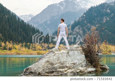 A man in white clothes with a sword stands on a large stone among the highlands. 71583642