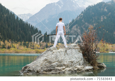 A man in white clothes with a sword stands on a large stone among the highlands. 71583643