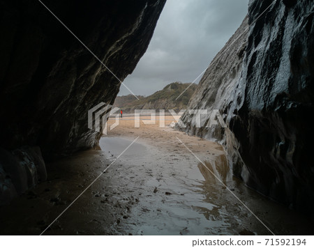 A small cave looking out to the sand Caswell Bay Beach in Gower, Wales, UK on a cloudy and wet day. A small cave looking out to the sand Caswell Bay Beach in Gower, Wales, UK on a cloudy and wet day. 71592194