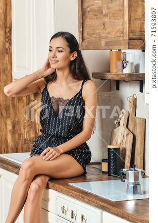 Smiling African American woman sitting on the kitchen counter and waiting for coffee to brew 71593807