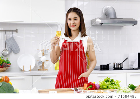 Happy Asian woman standing in the kitchen with glass of champagne. Concept woman cooking at home. Happy Asian woman standing in the kitchen with glass of champagne. Concept woman cooking at home. 71594485