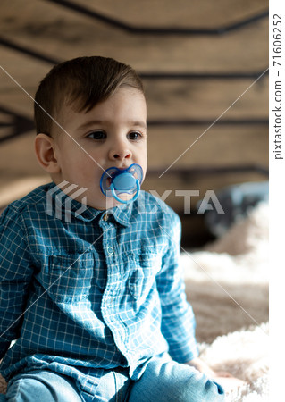 Childhood, family, loneliness concepts - Little preschool black boy in blue clothes Persian Arab appearance of two years of mixed nationality sitting on soft light bed with pacifier in mouth at home 71606252