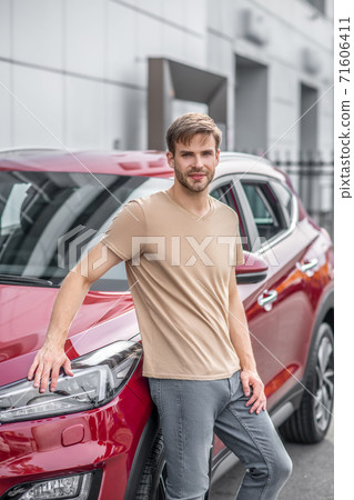 Smiling young male leaning on red car outside Smiling young male leaning on red car outside 71606411