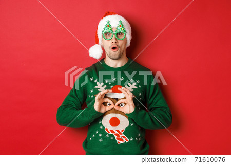 Portrait of carefree handsome man in santa hat and party glasses, making fun of his christmas sweater, looking happy over red background Portrait of carefree handsome man in santa hat and party glasses, making fun of his christmas sweater, looking happy over red background 71610076